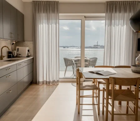Ljust kök och matplats i en modern hotellsvit med träbord, skandinavisk inredning och stora glasdörrar ut mot balkong med sittgrupp och sjöutsikt över glittrande vatten.