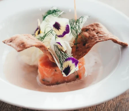 Förrätt med gravad lax, krispigt knäckebröd, färska örter, ätbara blommor och sås, serverad på vit tallrik.