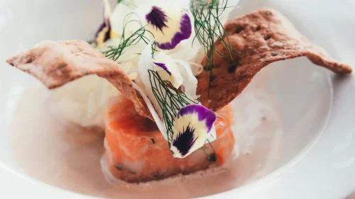 Förrätt med gravad lax, krispigt knäckebröd, färska örter, ätbara blommor och sås, serverad på vit tallrik.