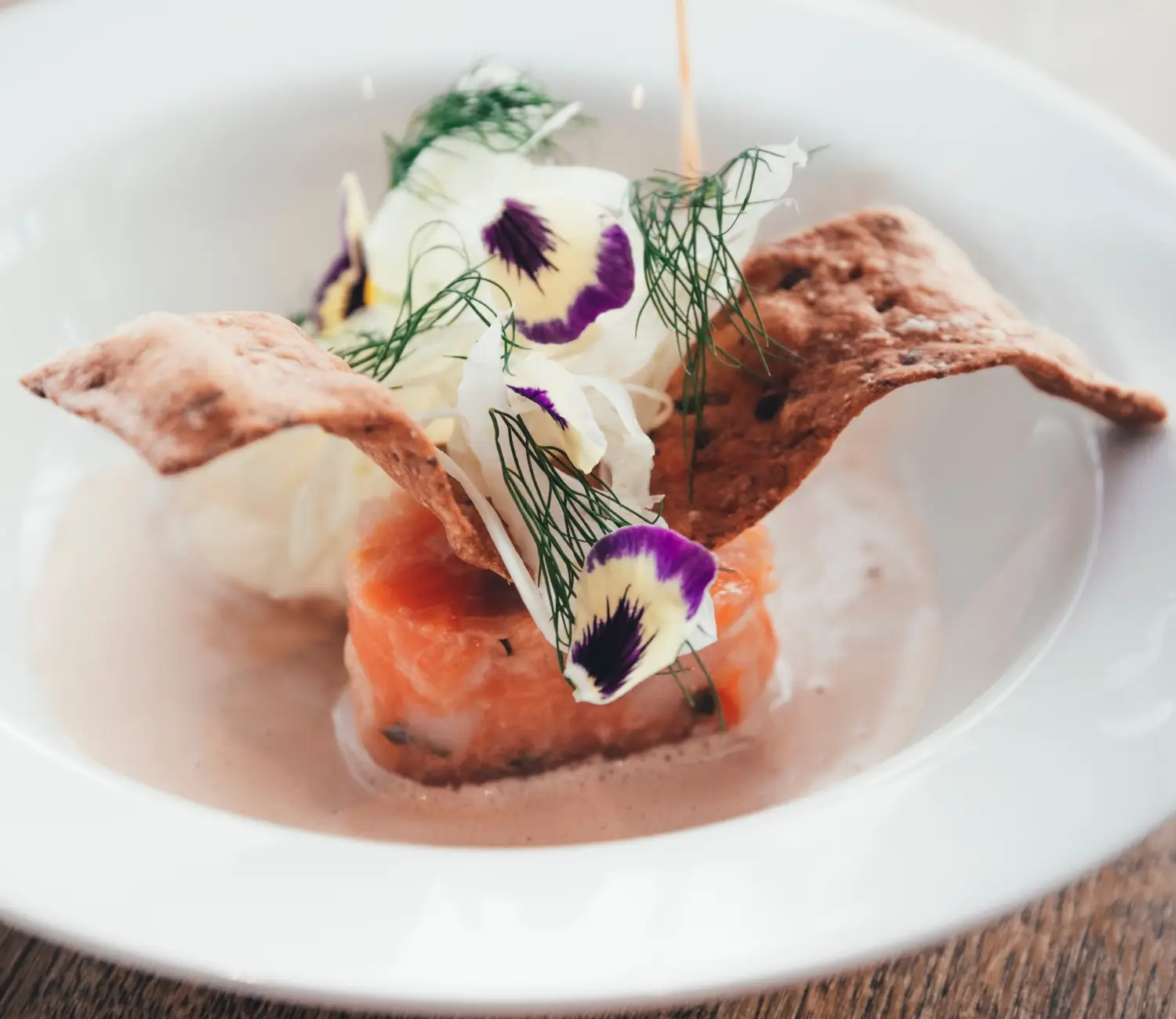 Förrätt med gravad lax, krispigt knäckebröd, färska örter, ätbara blommor och sås, serverad på vit tallrik.