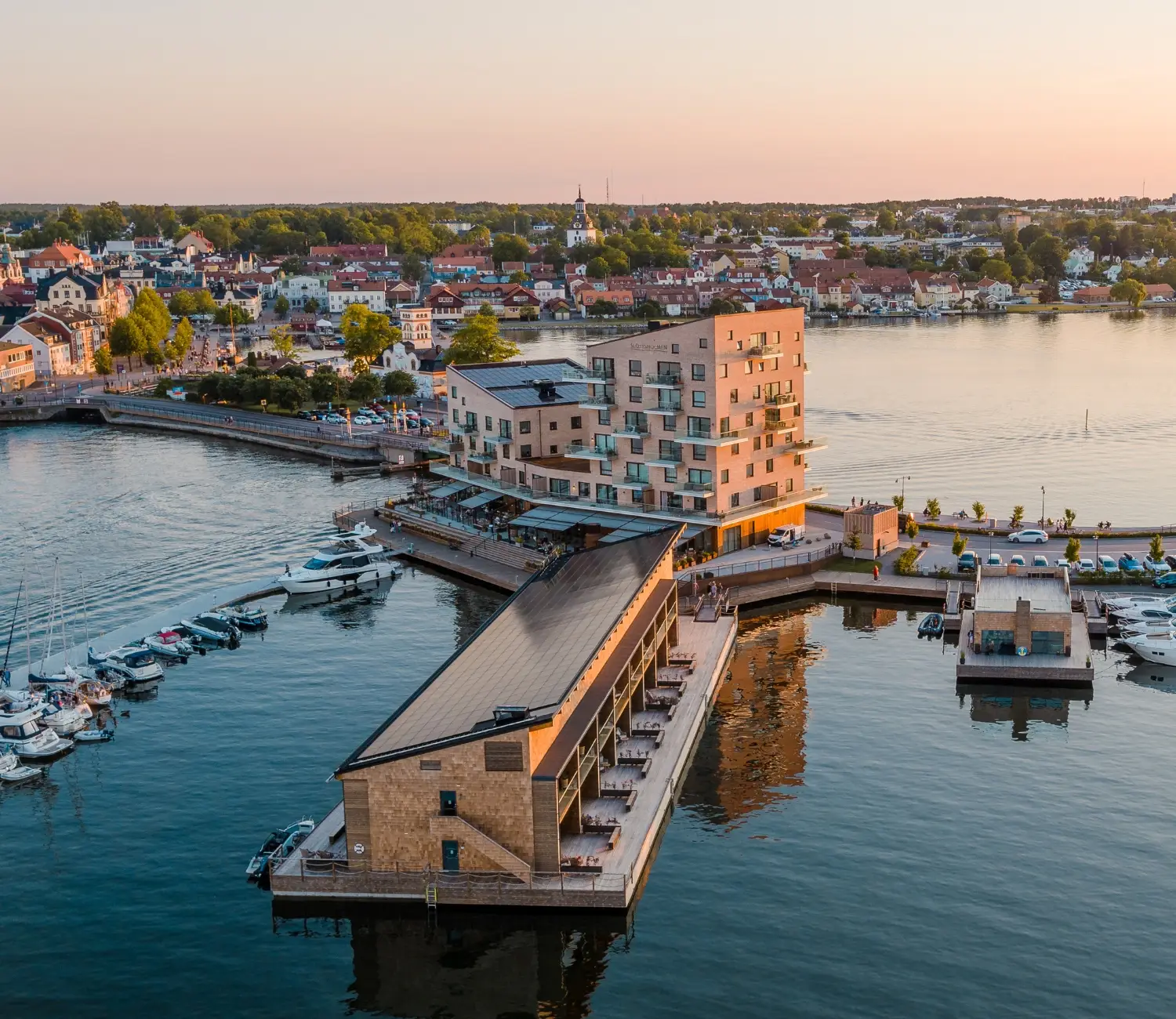Slottsholmen hotell i rosa kvällsljus med spegelblankt vatten runtom och Västervik i bakgrunden.