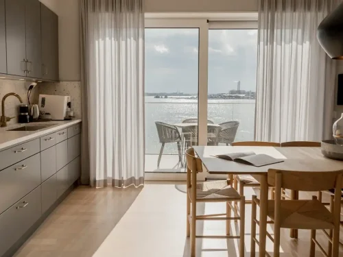 Ljust kök och matplats i en modern hotellsvit med träbord, skandinavisk inredning och stora glasdörrar ut mot balkong med sittgrupp och sjöutsikt över glittrande vatten.